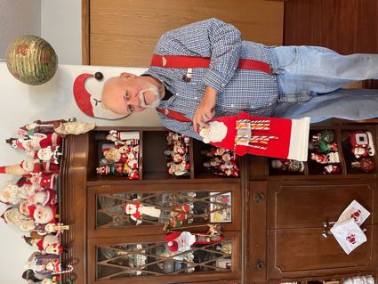 White man with white goatee stands in front of shelving with large number of Santa figurines.