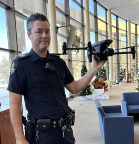 White male wearing a police uniform inside with a wall of windows behind him holding an unmanned aerial vehicle